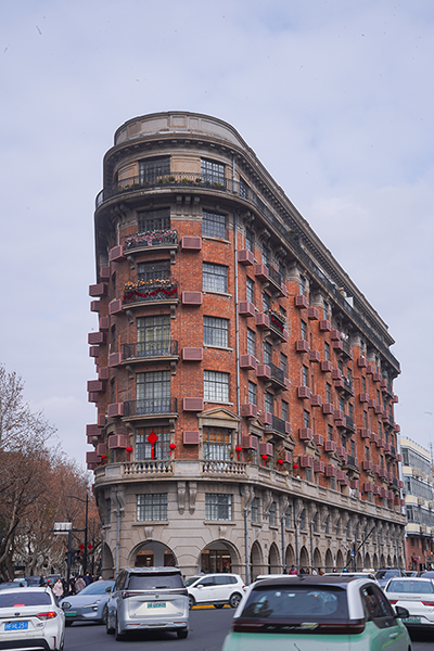 上海著名的武康大樓，一棟獨特的紅磚建築，外觀呈現弧形，設有多個陽台和綠色植物，周圍有許多汽車，背景為陰雲密布的天空。